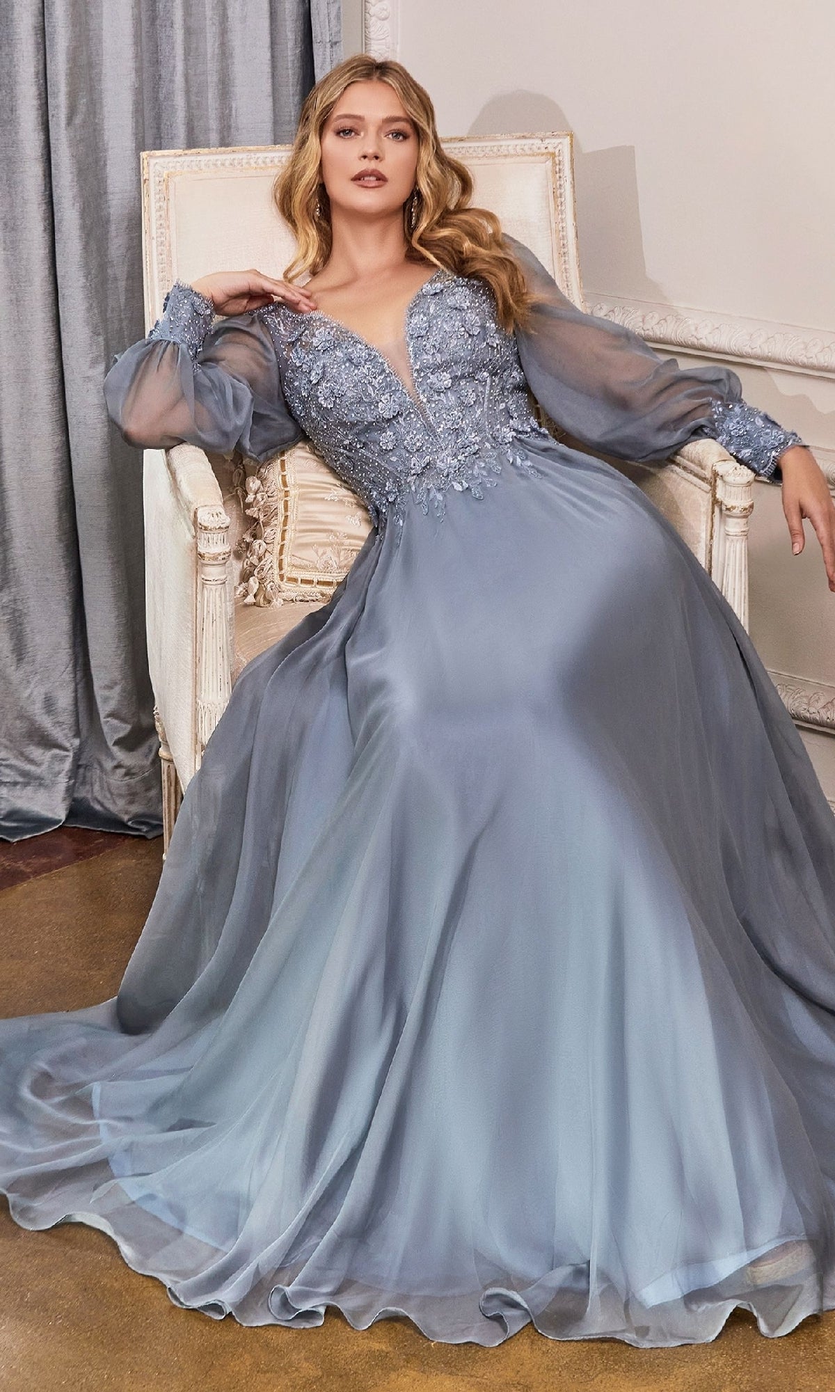 A woman wearing the Modest Long Sleeve Formal Gown: Ladivine CD0183, featuring sheer sleeves and floral embellishments, sits on a cream armchair.
